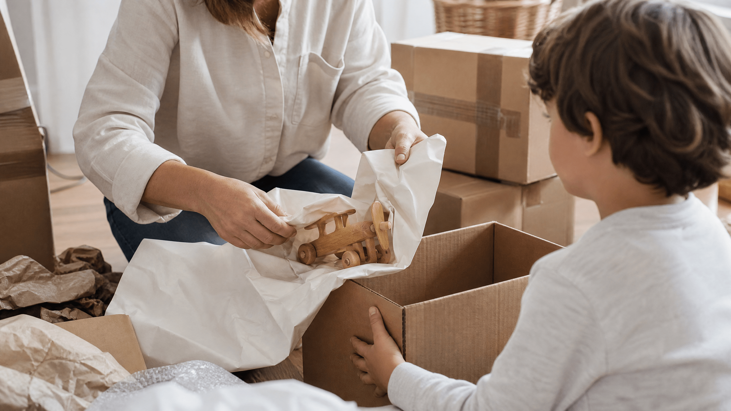 Kind und Elternteil schützen gemeinsam ein Holzspielzeug mit Papier für den Umzug.