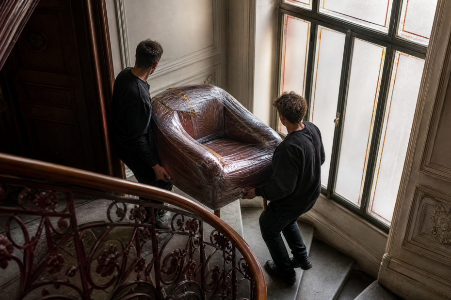 Zwei Umzugshelfer tragen einen geschützten Sessel durch ein historisches Berliner Altbautreppenhaus.